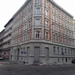 Odnowiona kamienica narożna z czerwoną cegłą i szarymi elementami dekoracyjnymi, widok z poziomu ulicy wyłożonej brukiem.