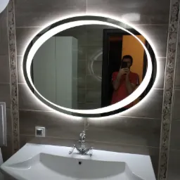Łazienka z umywalką i podświetlanym lustrem owalnym, widoczna osoba robiąca zdjęcie w odbiciu.