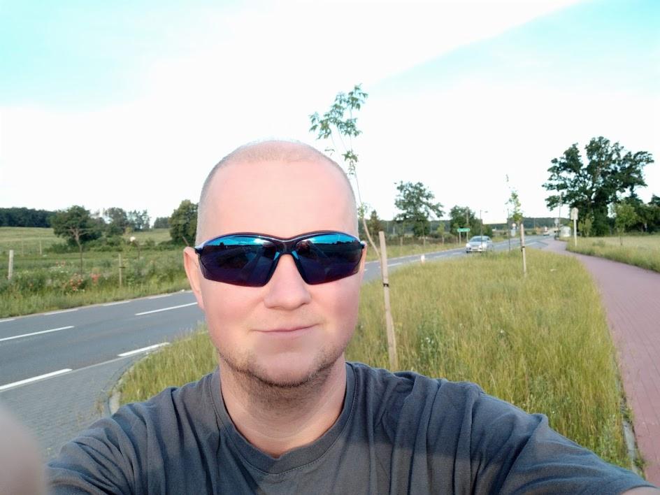 Mężczyzna w okularach przeciwsłonecznych robi selfie na tle drogi i zieleni. W tle widoczny samochód i drzewa, słoneczny dzień.