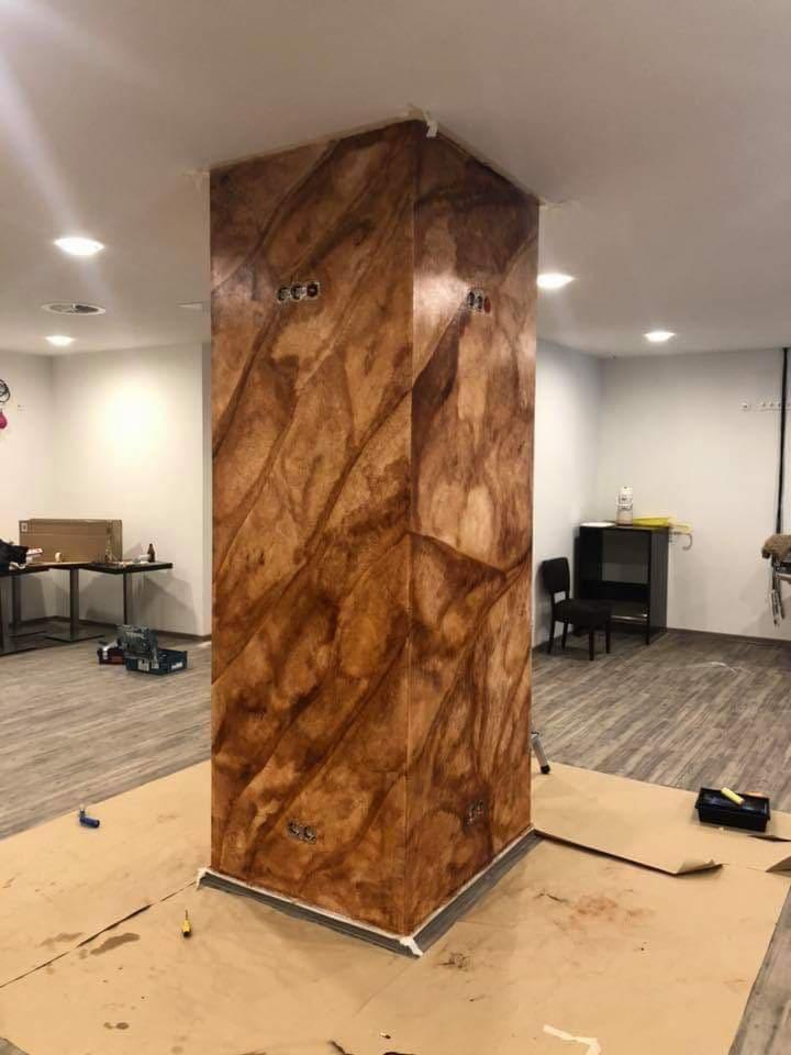 Kolumna w pomieszczeniu pokryta dekoracyjną, brązową farbą imitującą marmur, zabezpieczona kartonem na podłodze, widoczne gniazdka elektryczne i narzędzia malarskie.