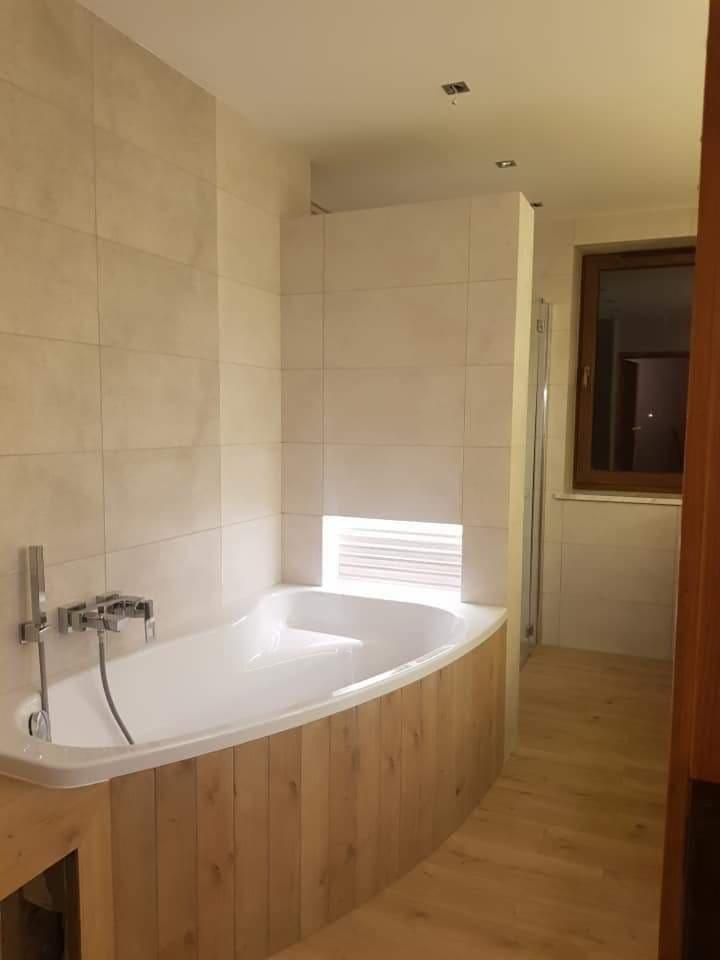 Wykończona łazienka z białą wanną obudowaną drewnianymi panelami, jasnymi płytkami na ścianach i podświetlaną wnęką nad wanną.