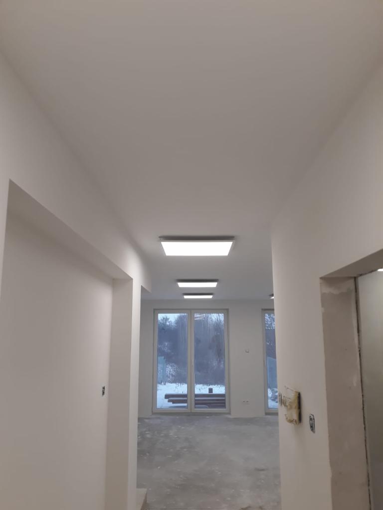 Wnętrze w trakcie remontu: widok z korytarza na pokój z dużymi oknami, świeżo pomalowane ściany na biało, sufit z zamontowanym oświetleniem LED, betonowa podłoga w stanie surowym.