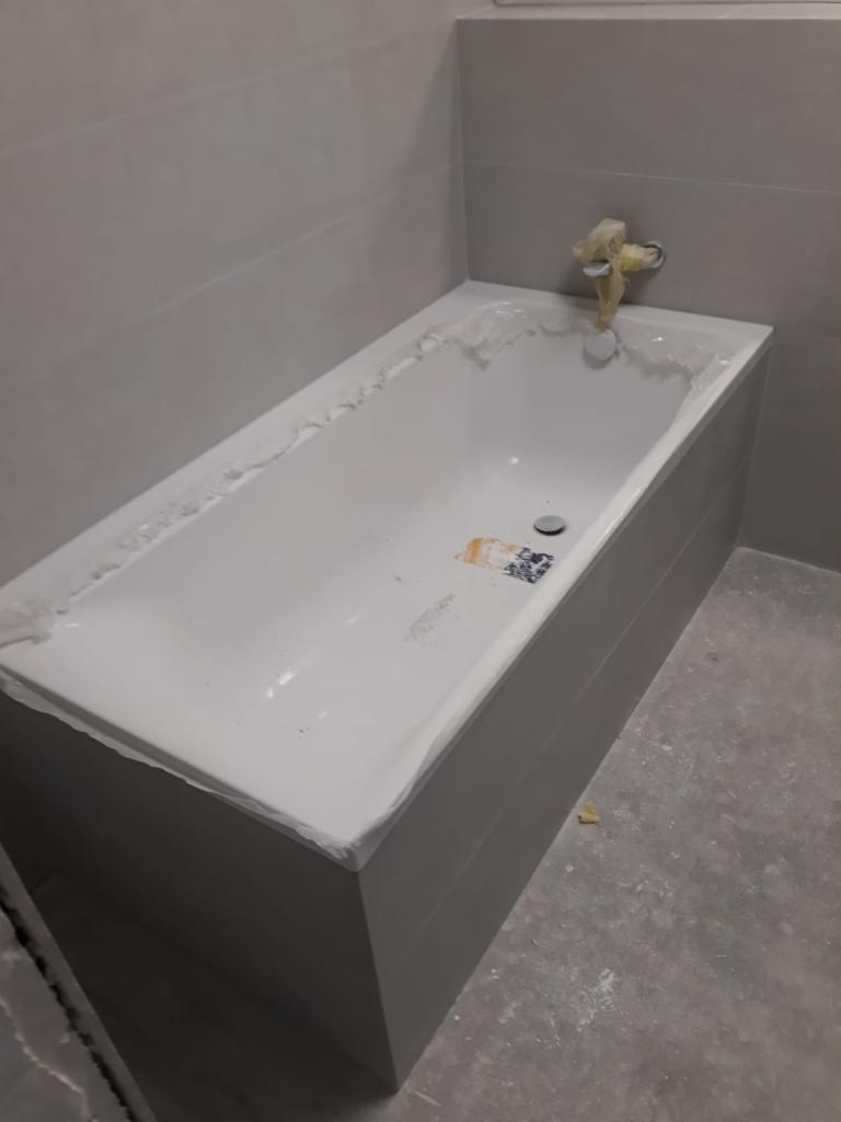Biała wanna z obudową w trakcie montażu, zabezpieczona folią malarską, widoczne ślady zaprawy na obudowie i podłodze, zakryta bateria.