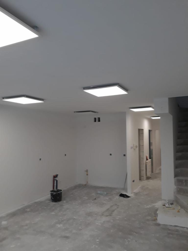 Wnętrze w trakcie remontu: świeżo pomalowane ściany na biało, widoczne kwadratowe panele LED w suficie, szpachlowane ściany z widocznymi punktami instalacji, wiadro farby i pozostałości po remoncie...