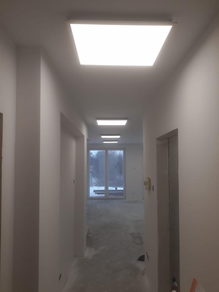 Korytarz w trakcie wykańczania z zamontowanymi panelami LED w suficie, widoczne ślady po pracach na ścianach i podłodze, okno z widokiem na zimowy krajobraz w tle.