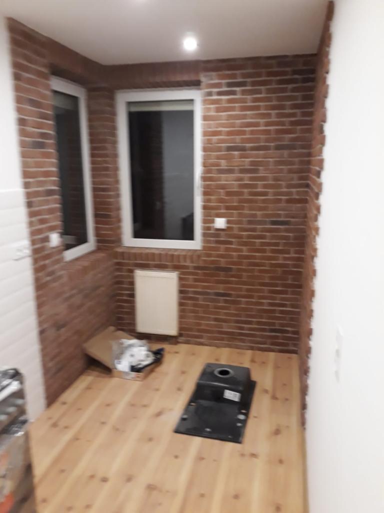 Wnętrze pomieszczenia z wykończonymi ścianami z cegły klinkierowej, drewnianą podłogą i oknami z białymi ramami; w rogu karton i czarny zlew przygotowany do montażu.