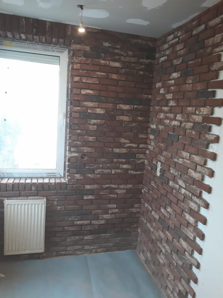 Pokój z ceglaną okładziną ścienną w odcieniach brązu i czerwieni, widoczny grzejnik pod oknem, sufit z widocznymi śladami po szpachlowaniu, w trakcie prac wykończeniowych.