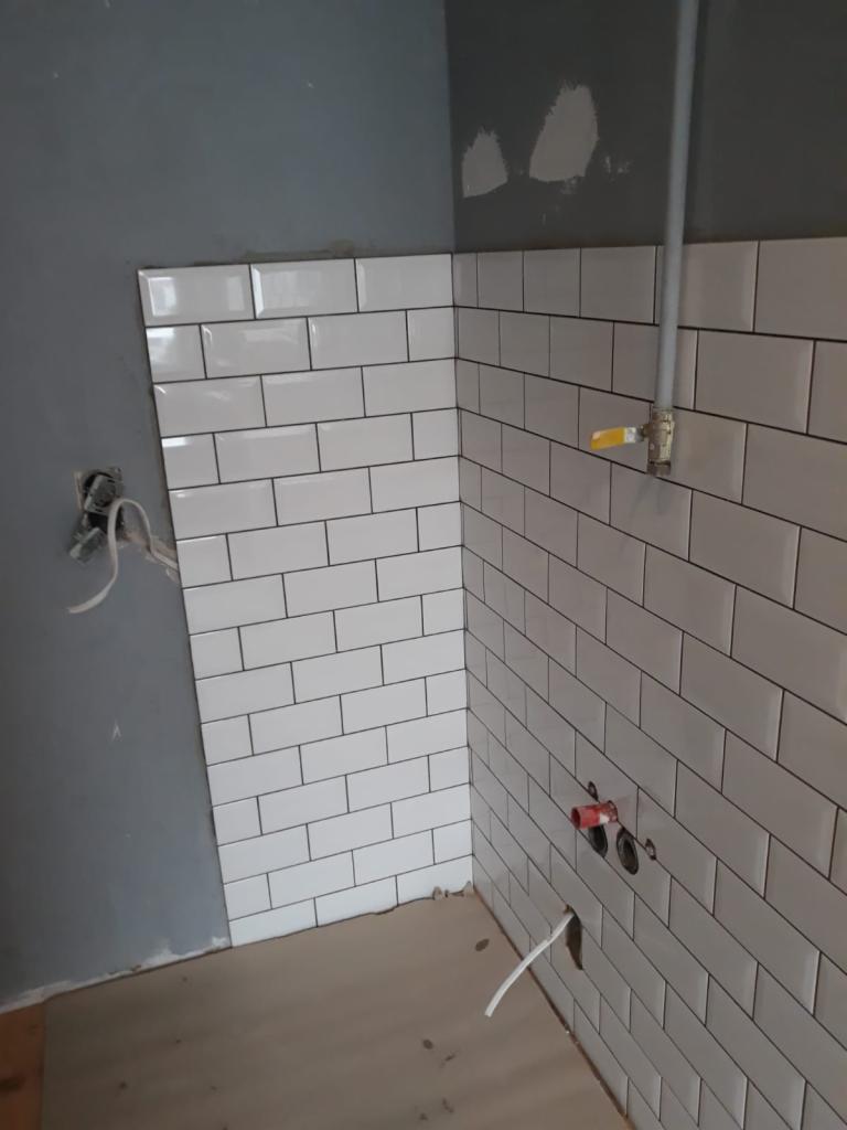 Świeżo położone białe, prostokątne płytki z czarną fugą na ścianie łazienki, widoczne wystające rury i kable, szara ściana obok częściowo wykończonej powierzchni.