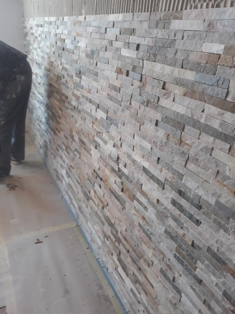 Ściana wyłożona dekoracyjnym kamieniem w odcieniach szarości, beżu i brązu, widoczne ślady zaprawy, fragment sylwetki osoby w roboczym ubraniu po lewej stronie.