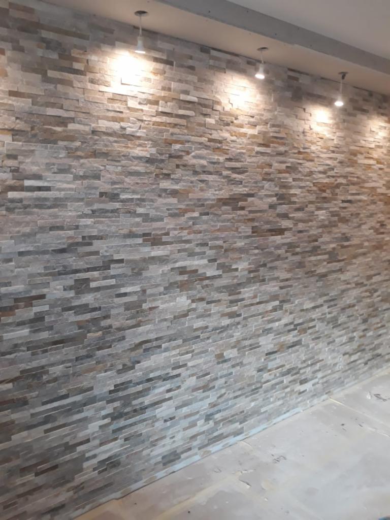 Ściana wykończona dekoracyjną okładziną kamienną w odcieniach szarości i brązu, oświetlona punktowymi lampami sufitowymi, widoczne niedoskonałości fugowania w dolnej części ściany.