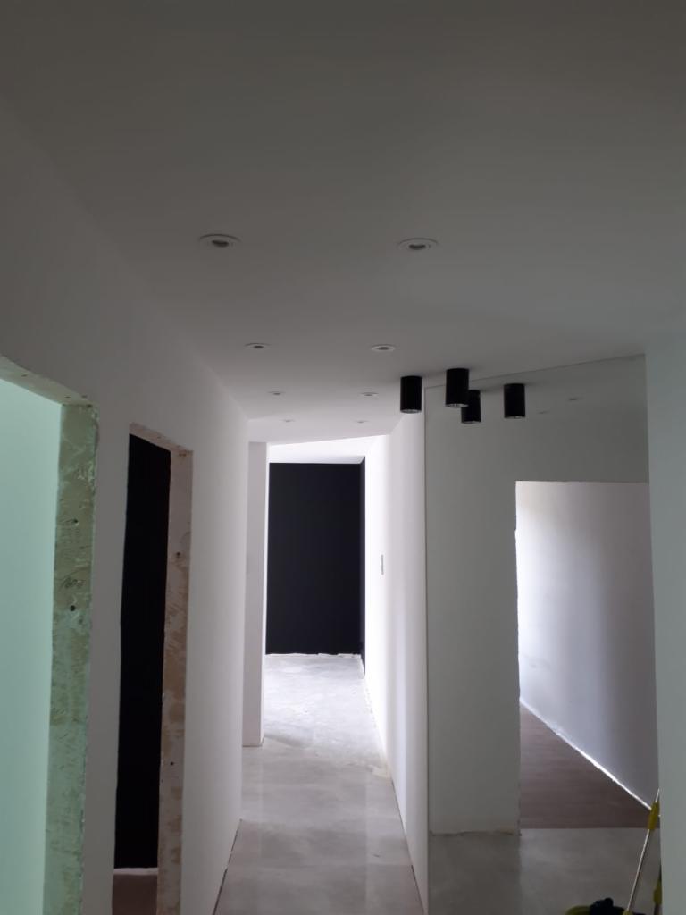 Korytarz w trakcie remontu z widocznymi surowymi ścianami, nowymi czarnymi lampami sufitowymi i czarną ścianą w oddali, ukazujący postęp prac wykończeniowych.