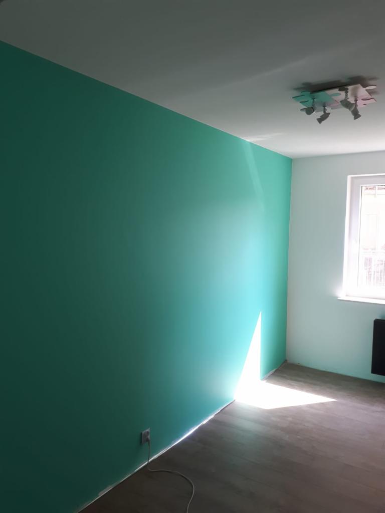 Świeżo pomalowane wnętrze pokoju z jedną ścianą w kolorze turkusowym, kontrastującą z jasną ścianą obok okna, drewniana podłoga, widoczne gniazdko elektryczne i nowoczesna lampa sufitowa.