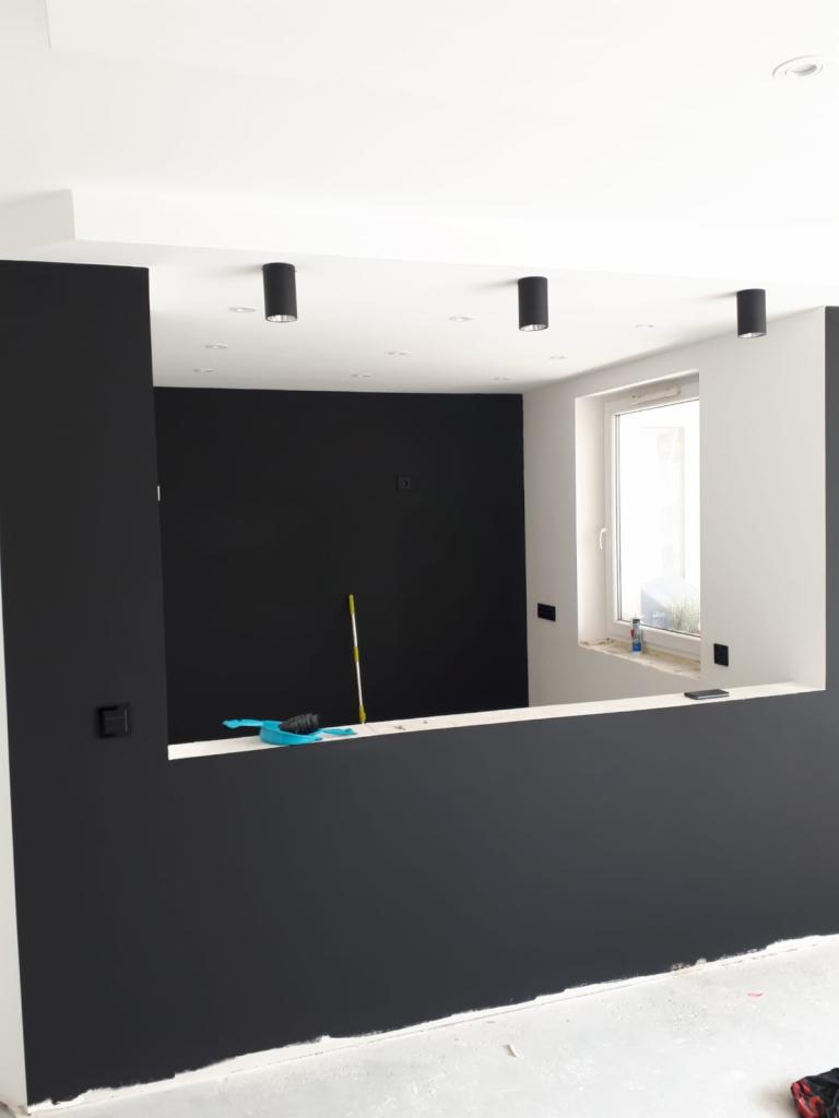 Wnętrze w trakcie malowania na czarno, widoczne narzędzia malarskie, surowa podłoga i okno z białymi ramami.