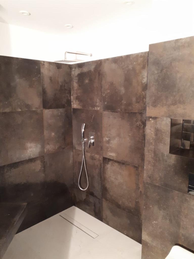 Nowoczesna kabina prysznicowa z ciemnymi, metalicznymi płytkami o nieregularnej fakturze, wyposażona w deszczownicę i słuchawkę prysznicową, z odpływem liniowym w białej posadzce.
