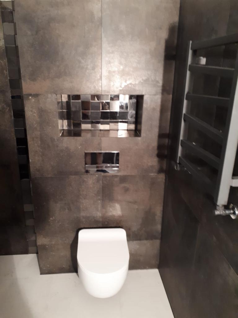 Nowoczesna toaleta podwieszana z mozaiką wnękową i grzejnikiem drabinkowym na tle płytek imitujących metal.