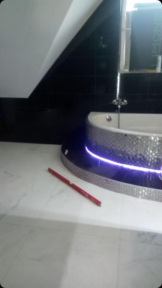 Nowoczesna łazienka w trakcie wykańczania: czarne płytki ścienne, wanna z mozaiką i oświetleniem LED, poziomica na marmurowej podłodze.