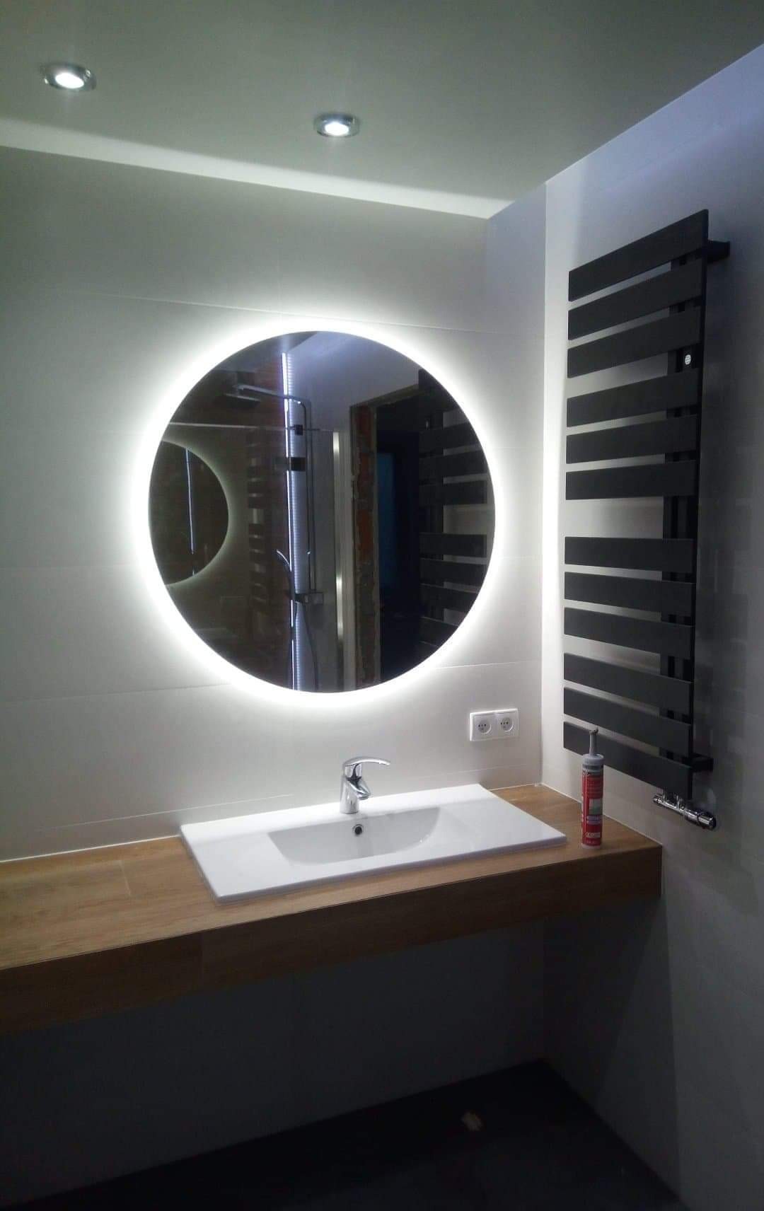 Wykończona łazienka z okrągłym lustrem LED, drewnianym blatem pod umywalką, grzejnikiem drabinkowym w kolorze czarnym i białymi płytkami na ścianach. Widoczna tuba silikonu sanitarnym obok umywalki.