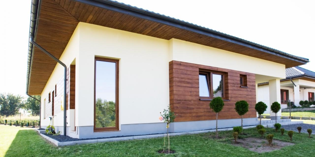 Nowoczesny dom jednorodzinny z drewnianą elewacją, brązowymi oknami i dachem, otoczony zielonym trawnikiem i młodymi drzewkami.