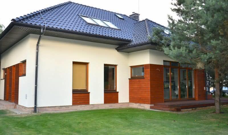 Nowoczesny dom jednorodzinny z ciemną dachówką i oknami dachowymi, wykończony drewnem na elewacji, otoczony zielonym trawnikiem.