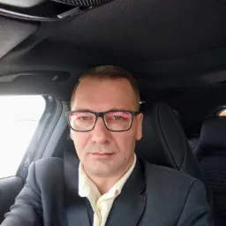 Mężczyzna w garniturze i okularach siedzący w samochodzie, prawdopodobnie w drodze na spotkanie z klientem.