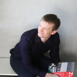 Uśmiechnięty mężczyzna w ciemnej koszuli, siedzący na białym grzejniku, trzymający w rękach katalog z projektami wnętrz i elementami graficznymi.