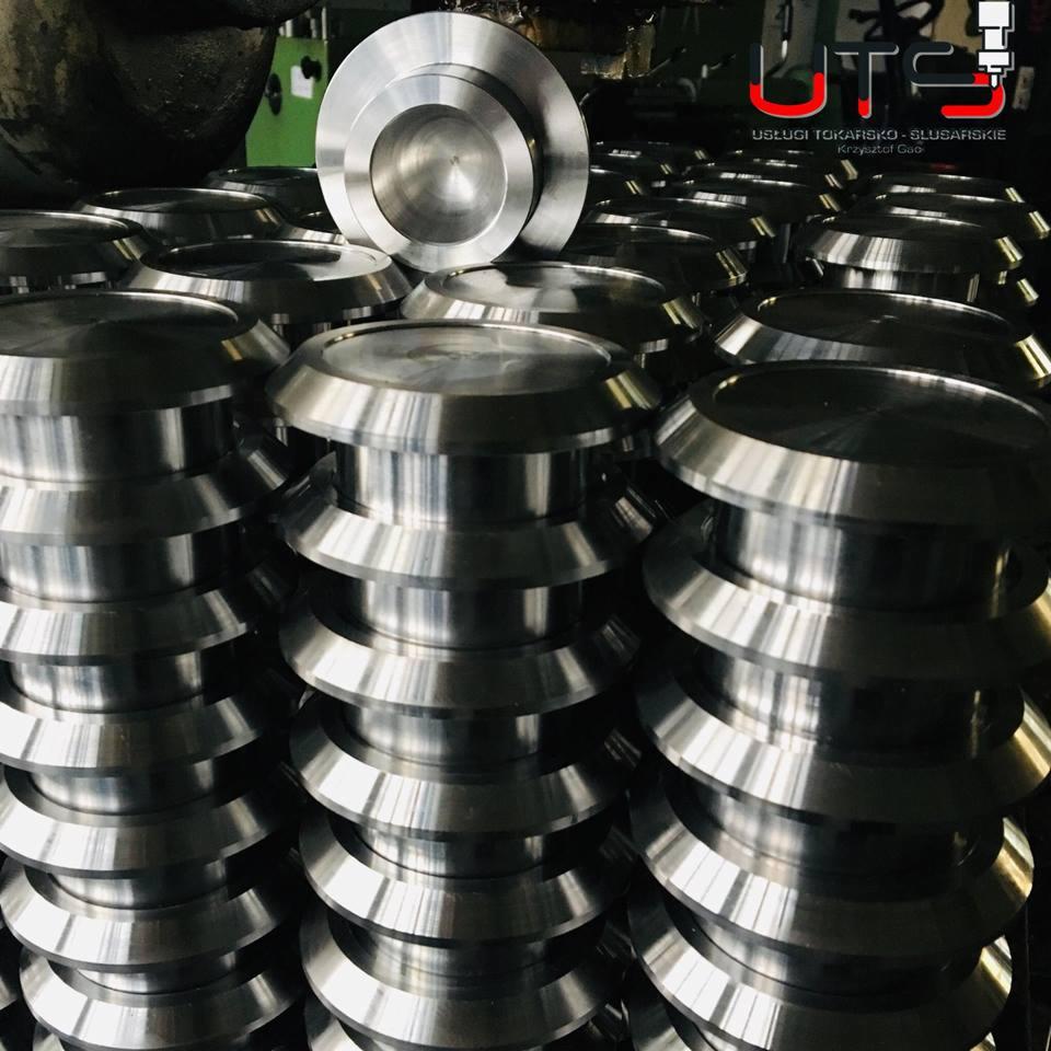 Stos metalowych, obrobionych elementów o okrągłym kształcie z widocznymi śladami obróbki, ułożonych jeden na drugim, z logo firmy w tle.