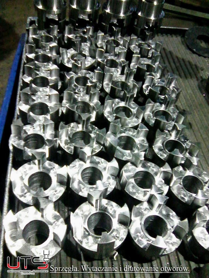 Partia precyzyjnie obrobionych, metalowych elementów z centralnym otworem i zewnętrznymi wypustkami, ułożonych w niebieskiej skrzynce, widoczne ślady obróbki skrawaniem.