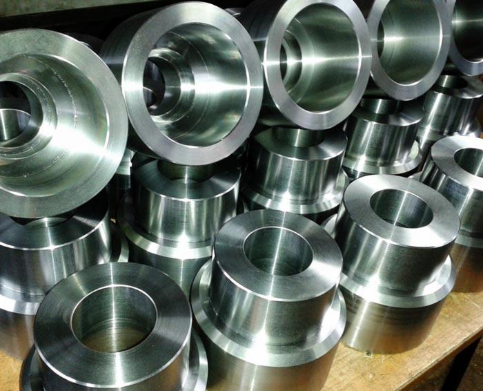 Stos obrobionych, metalowych tulei o różnych średnicach, ułożonych warstwami na drewnianej powierzchni, z widocznymi śladami obróbki i refleksami światła.