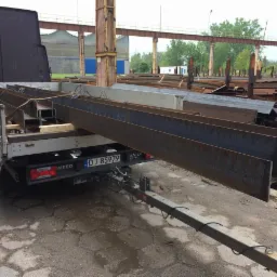 Płaski samochód dostawczy Iveco z przyczepą przewozi stalowe profile konstrukcyjne typu I, na tle hali przemysłowej.