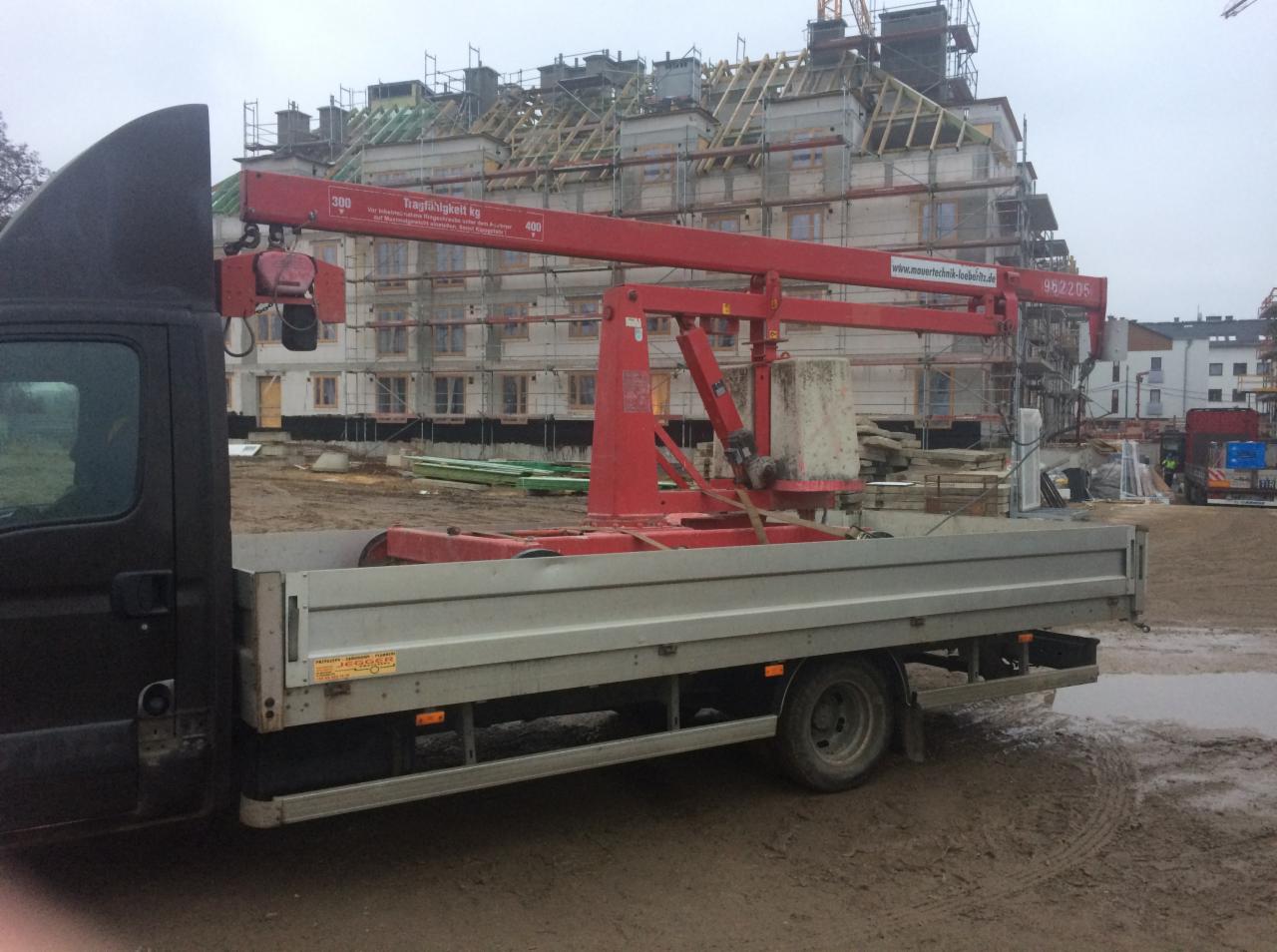 Czarny bus z czerwoną platformą dźwigową na pace, na tle budynku w trakcie budowy, widoczne rusztowania i dźwig budowlany.