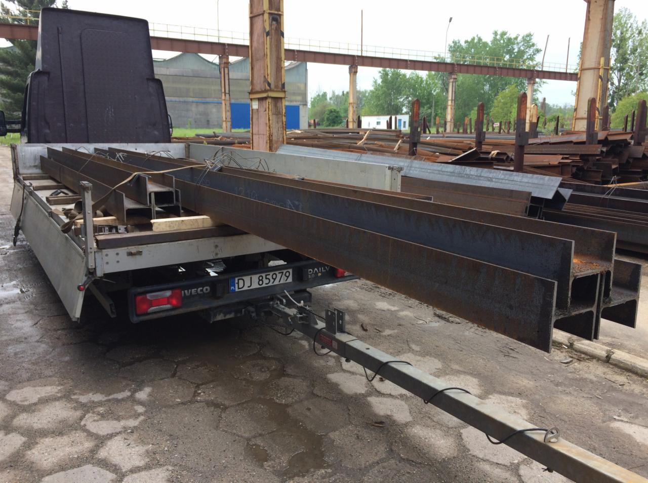 Płaski samochód dostawczy Iveco z przyczepą przewozi stalowe profile konstrukcyjne typu I, na tle hali przemysłowej.