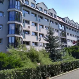 Wielopiętrowy budynek mieszkalny z szarą elewacją, balkonami z przeszkleniami i zadbaną zielenią w otoczeniu.