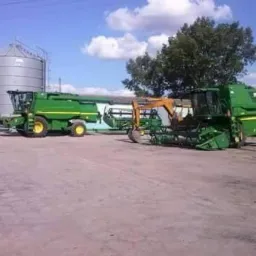 Dwie zielone, nowoczesne maszyny rolnicze stoją na placu obok silosów zbożowych w słoneczny dzień. Widoczny fragment koparki.