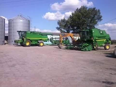 Dwie zielone, nowoczesne maszyny rolnicze stoją na placu obok silosów zbożowych w słoneczny dzień. Widoczny fragment koparki.