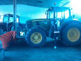 Zielony traktor John Deere z żółtymi felgami, widoczny w hali, z pomarańczową rurą na pierwszym planie i drugim traktorem w tle. Oświetlenie hali tworzy niebieską poświatę.