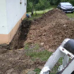Wykop wzdłuż fundamentu budynku, usypana pryzma ziemi, widoczny fragment maszyny budowlanej i zaparkowany samochód osobowy w tle.