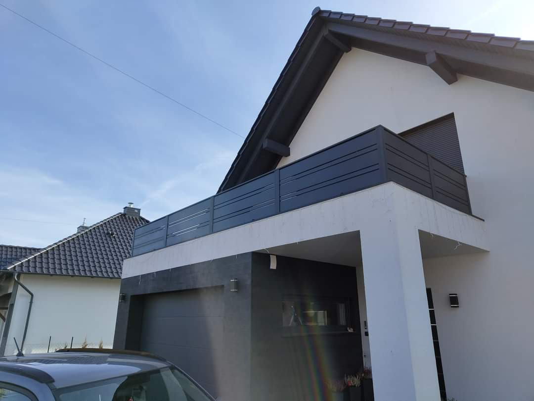 Nowoczesny balkon z szarą, poziomą balustradą zamontowany przy domu z szarą stolarką okienną i ciemną dachówką, widok z dołu na jasnym tle nieba.