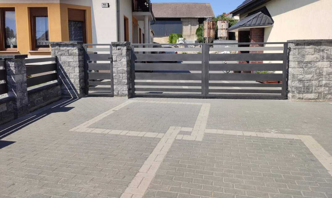 Nowoczesna, pozioma brama przesuwna i furtka w kolorze grafitowym, z kamiennymi słupkami, na posesji z kostką brukową w odcieniach szarości.