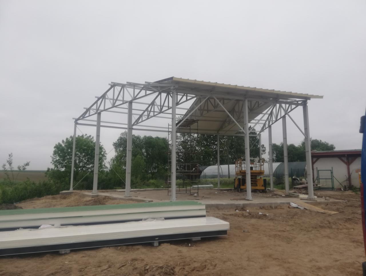 Konstrukcja stalowa hali w trakcie budowy, widoczne elementy konstrukcyjne, częściowo zamontowane panele dachowe, podnośnik koszowy i stos płyt warstwowych na ziemi.