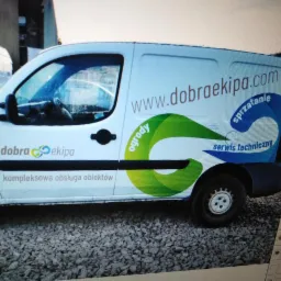 Biały samochód dostawczy z logo firmy 'dobra ekipa' oferującej kompleksową obsługę obiektów, w tym usługi ogrodnicze, sprzątanie i serwis techniczny, zaparkowany na żwirowym podłożu.