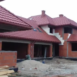 Niedokończony dom jednorodzinny z czerwonym dachem, widoczne ściany z pustaków ceramicznych, brak okien i drzwi, zarys schodów zewnętrznych, na placu budowy piasek i materiały budowlane.