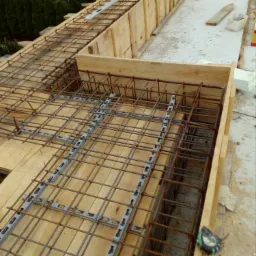 Zbrojenie fundamentu w drewnianym szalunku, widoczne elementy dystansowe z tworzywa sztucznego i taśma miernicza przy krawędzi szalunku.