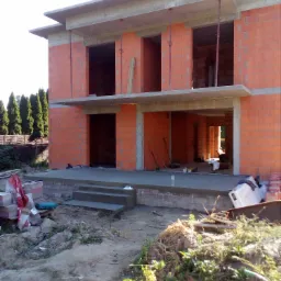 Surowy stan dwukondygnacyjnego domu z czerwonej cegły z widocznymi otworami okiennymi i betonowym stropem, wspartego na stalowych podporach; widoczne schody wejściowe i materiały budowlane na placu...