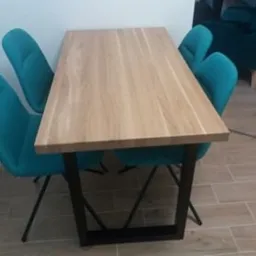 Stół z blatem z jasnego drewna i czarnymi, metalowymi nogami w kształcie trapezu. Cztery turkusowe krzesła z tapicerowanym siedziskiem i oparciem otaczają stół. W tle widoczny fragment turkusowego...