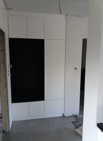 Nowoczesna szafa wnękowa z białymi frontami i czarnym panelem, zamontowana w surowym wnętrzu z widocznymi niedokończonymi pracami wykończeniowymi.