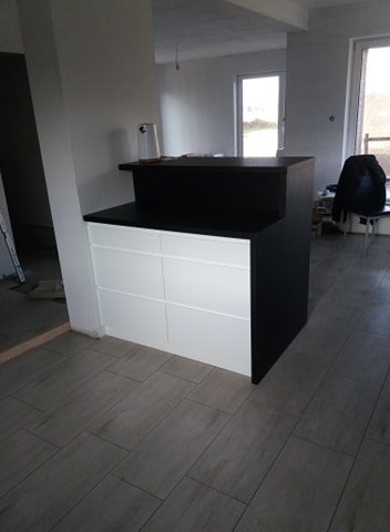 Wyspa kuchenna z białymi frontami szafek i czarnym blatem w surowym wnętrzu, widok z boku na podłogę z płytek imitujących drewno.