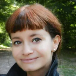 Portret kobiety z brązowymi oczami i kasztanowymi włosami z grzywką na tle zielonego parku.