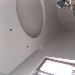 Firma Remontowo Budowlana Tomasz Piechota - Biały sufit podwieszany z okrągłym wgłębieniem i punktowymi otworami na oświetlenie, widoczne lampy halogenowe i puszki z pianką montażową przy oknie.