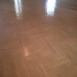 Nowo położona, lakierowana parkietowa podłoga ułożona we wzór jodełki francuskiej, odbijająca światło z okna w pustym pomieszczeniu.