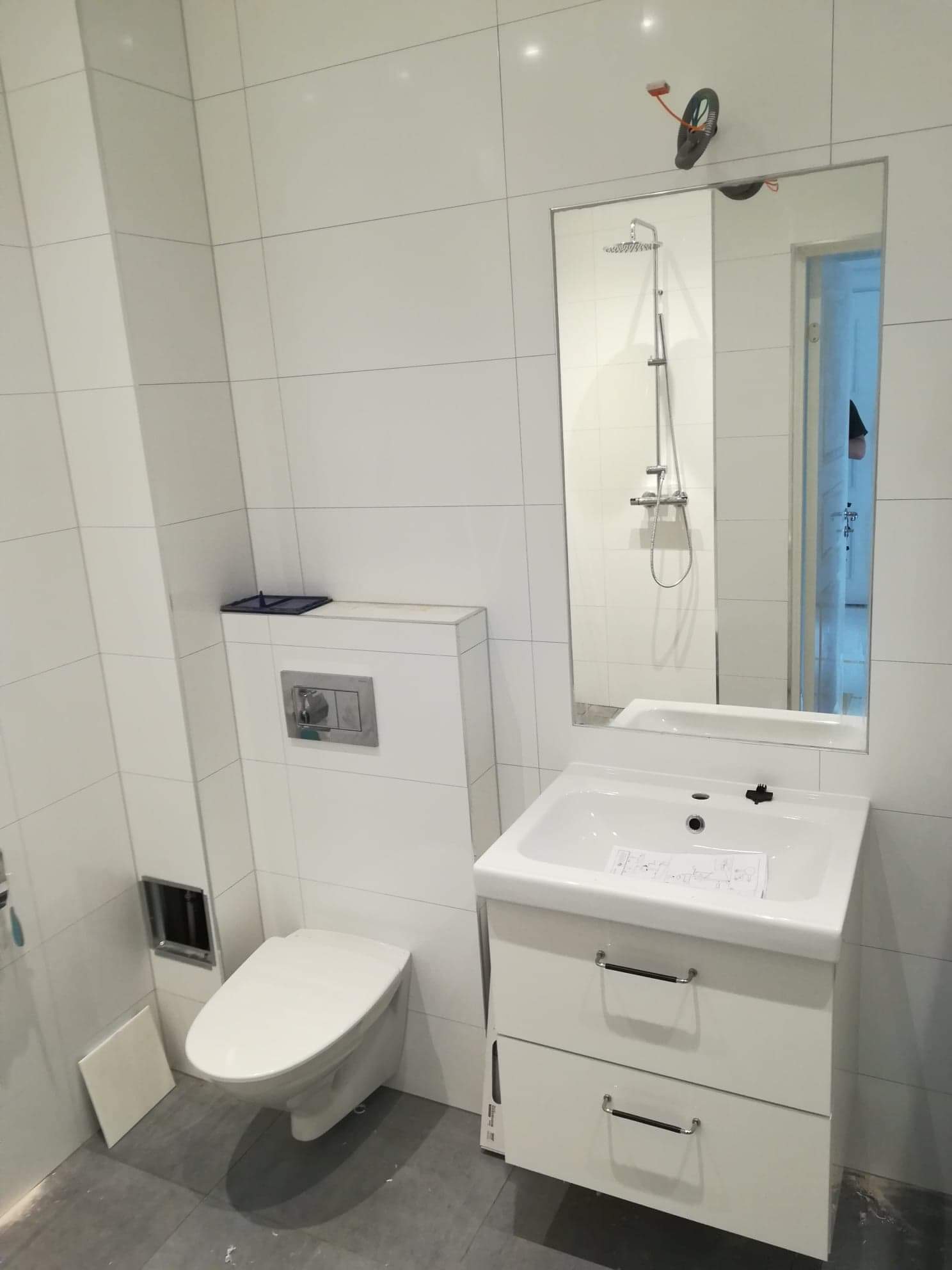 Nowoczesna łazienka z białymi płytkami, wiszącą toaletą i szafką z umywalką. Na umywalce leżą plany lub schematy. W lustrze odbija się prysznic.